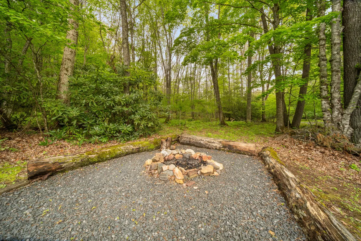 A cozy stone fire pit surrounded by rustic log seating sits nestled in a lush, green yard. The area is bordered by dense trees, creating a secluded atmosphere ideal for evening gatherings, marshmallow roasting, and storytelling under the stars.