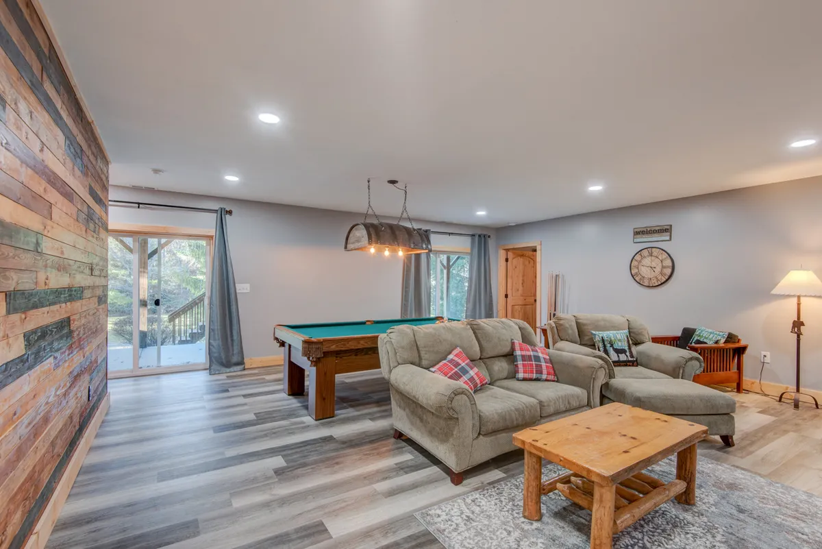 A spacious game room featuring a pool table in the center, comfortable seating, a wooden accent wall, and a warm lighting fixture overhead. The arrangement encourages group activities and social gatherings, evoking a sense of fun and friendly competition.