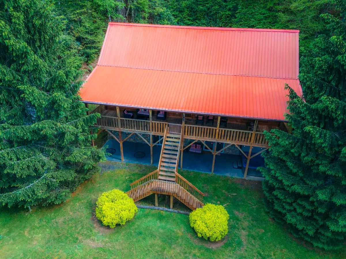 This image showcases the cabin's red metal roof, wooden staircase, and spacious deck area, set amidst lush greenery. The inviting deck provides a glimpse of the mountain environment and outdoor gathering spaces ideal for group relaxation and meals.