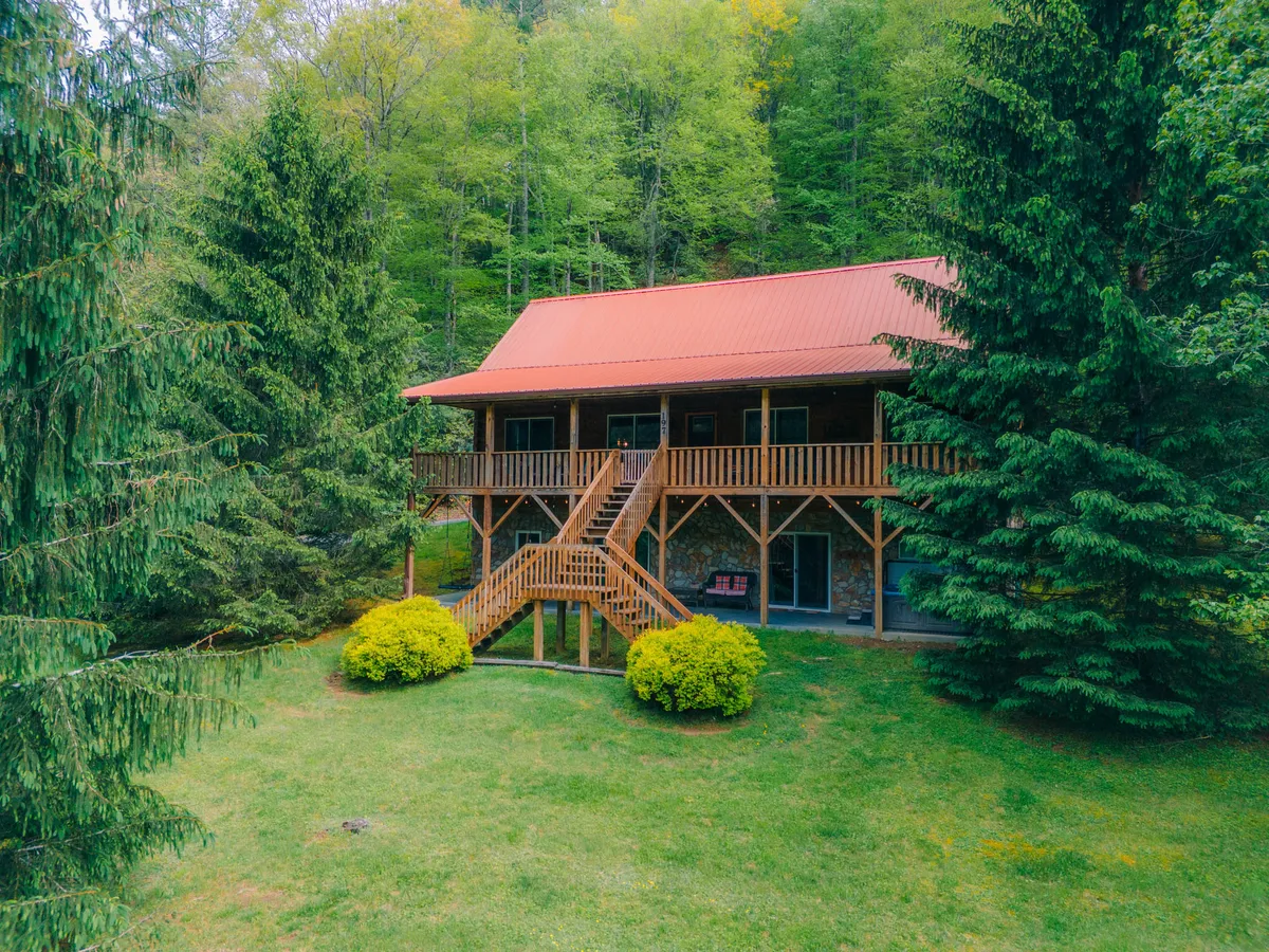 The image showcases a charming log cabin with a striking red metal roof and a spacious wooden deck, set against a backdrop of lush, verdant trees. The stone foundation and cozy porch invite relaxation, while the surrounding greenery hints at privacy and immersion in nature. The scene exudes tranquility, perfect for unwinding or gathering with loved ones outdoors.