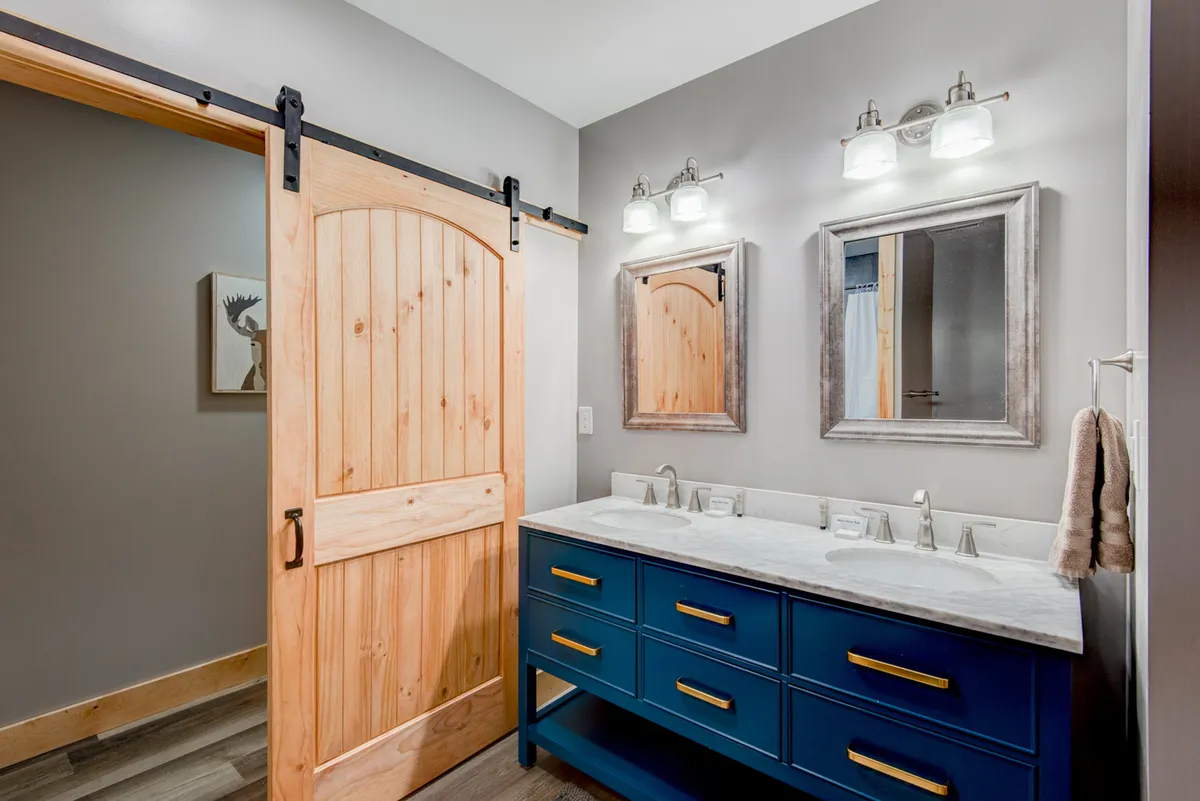 A spacious bathroom with double vanity sinks, mirrors, and soft lighting fixtures set against a calming backdrop. The barn door adds a touch of rustic charm, and the overall mood is serene and elegant.