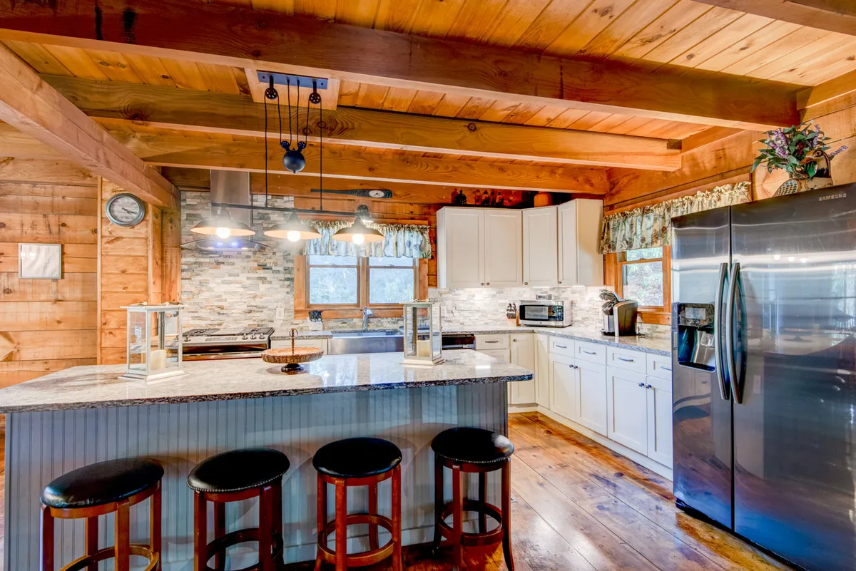 A close-up of a kitchen island with granite countertops, stainless steel appliances, and bar stools, framed by rustic wooden beams and a stone backsplash. The inviting setup reflects a hub for cooking, entertaining, and casual breakfasts.