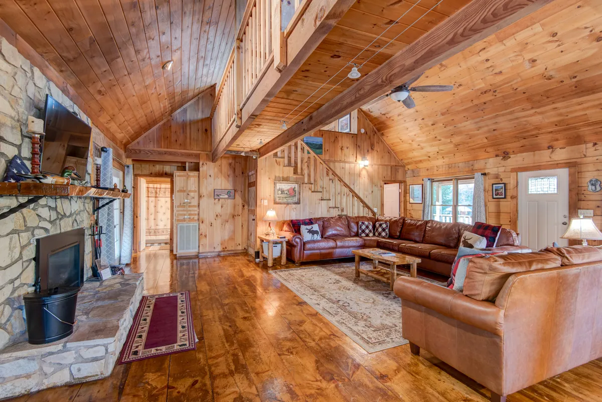 This inviting scene features a great room with a stone fireplace as its centerpiece, surrounded by a large leather sofa and accented by sturdy wooden beams and a staircase. The setup reflects a warm, communal environment ideal for family gatherings, storytelling, and evenings by the fire.