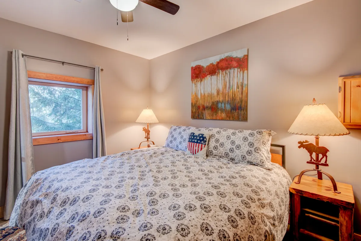 A serene bedroom featuring a bed with patterned bedding, artwork on the wall, table lamps, and wooden furniture. The natural light filtering into the room accentuates the tranquil and inviting atmosphere, suggesting a restful space ideal for a peaceful night's sleep.