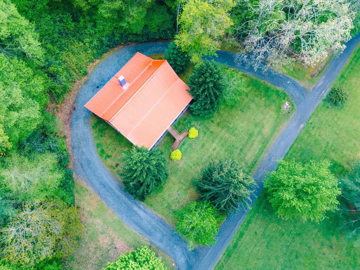 This exterior image highlights the cabin with its distinctive red roof, surrounded by lush greenery and a circular driveway. The scene conveys a peaceful, welcoming mountain retreat, with open space ideal for outdoor activities and gentle yard slopes visible around the property.