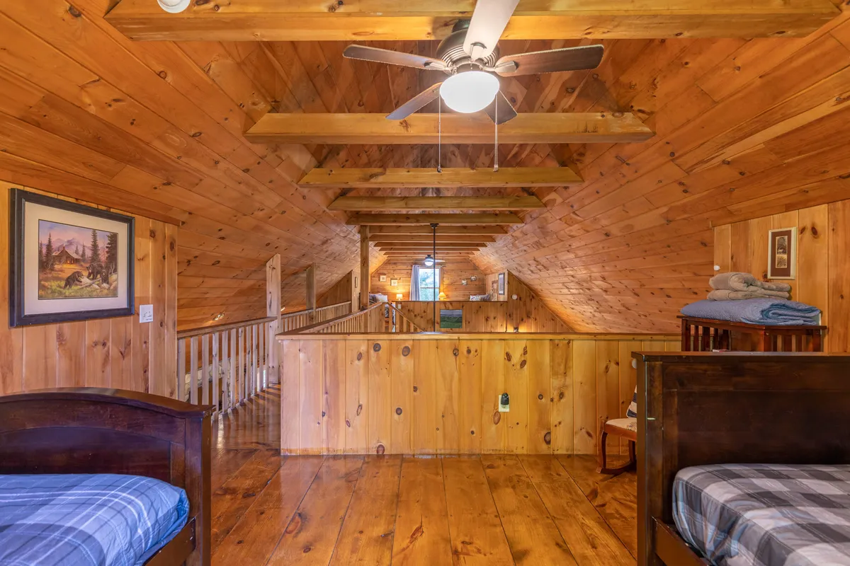 An open loft space featuring wooden beams, a ceiling fan, bunk beds, framed artwork, and a wooden railing. The open design suggests a connection to other loft spaces, and the cozy furnishings make it ideal for both children and adults to relax or play.