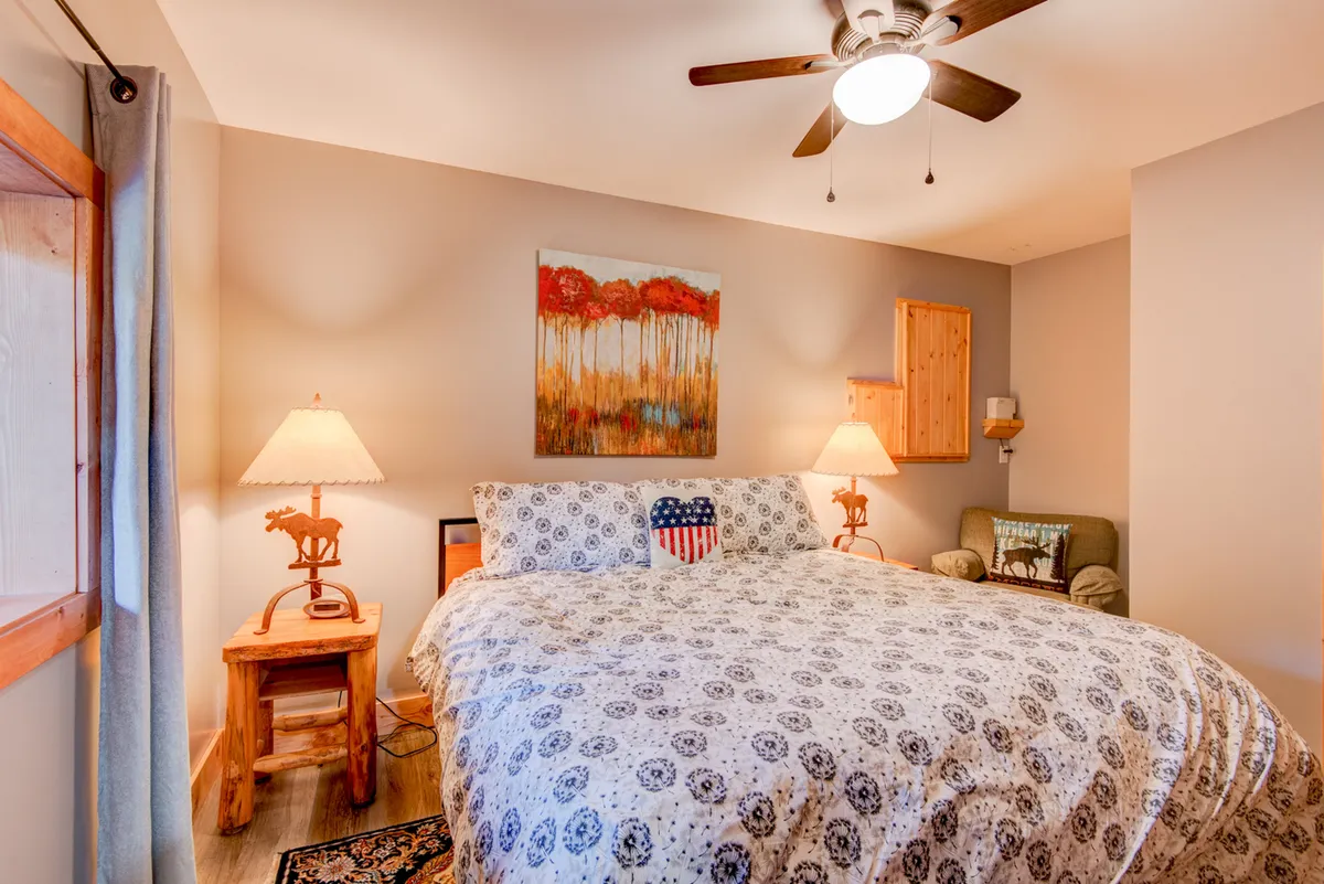 A cozy bedroom with a bed featuring patterned bedding, a wooden nightstand, table lamps, and artwork on the wall. The overall feel is warm and inviting, ideal for relaxing, reading, or sleeping in comfort.